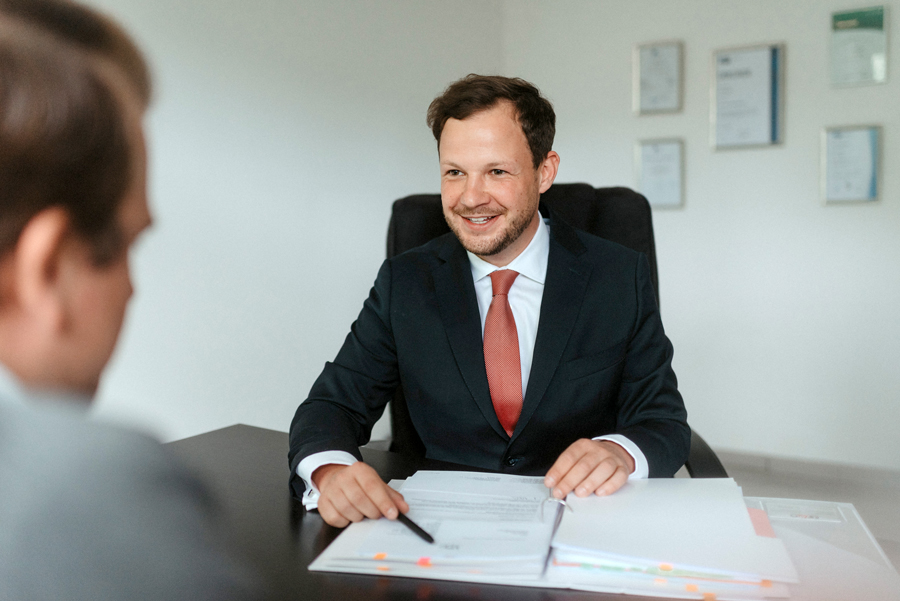Ein Mann im Anzug und mit roter Krawatte sitzt an einem Schreibtisch, lächelt und hält einen Stift in der Hand, während er mit einer anderen Person spricht. Auf dem Schreibtisch sind Dokumente und Ordner ausgebreitet, und an der Wand hinter ihm hängen Urkunden.