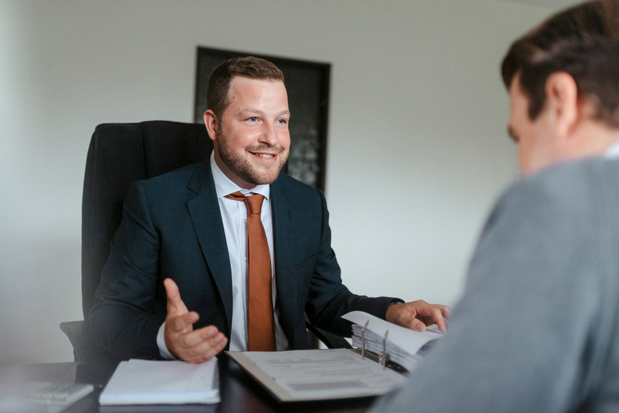 Ein Mann im Anzug sitzt an einem Schreibtisch, lächelt und spricht mit einer anderen Person. Sie sind von Dokumenten und Ordnern umgeben, was auf eine berufliche Besprechung oder ein Gespräch in einem Büro schließen lässt.